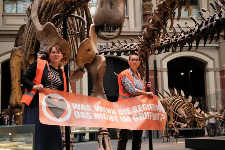 Las dos ambientalistas pegadas a barras metálicas alrededor de una exposición de esqueletos de dinosaurios y sosteniendo una pancarta. Foto: AFP.
