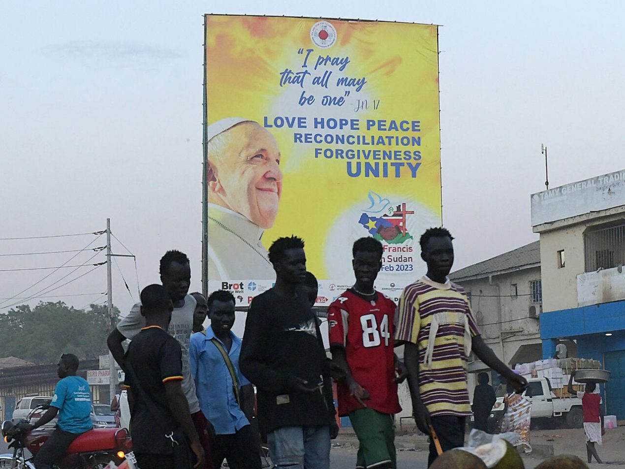 Un grupo de jóvenes camina en Juba cerca de un cartel alusivo a la visita del papa Francisco a Sudán del Sur el 31 de enero de 2023. Foto: AFP.